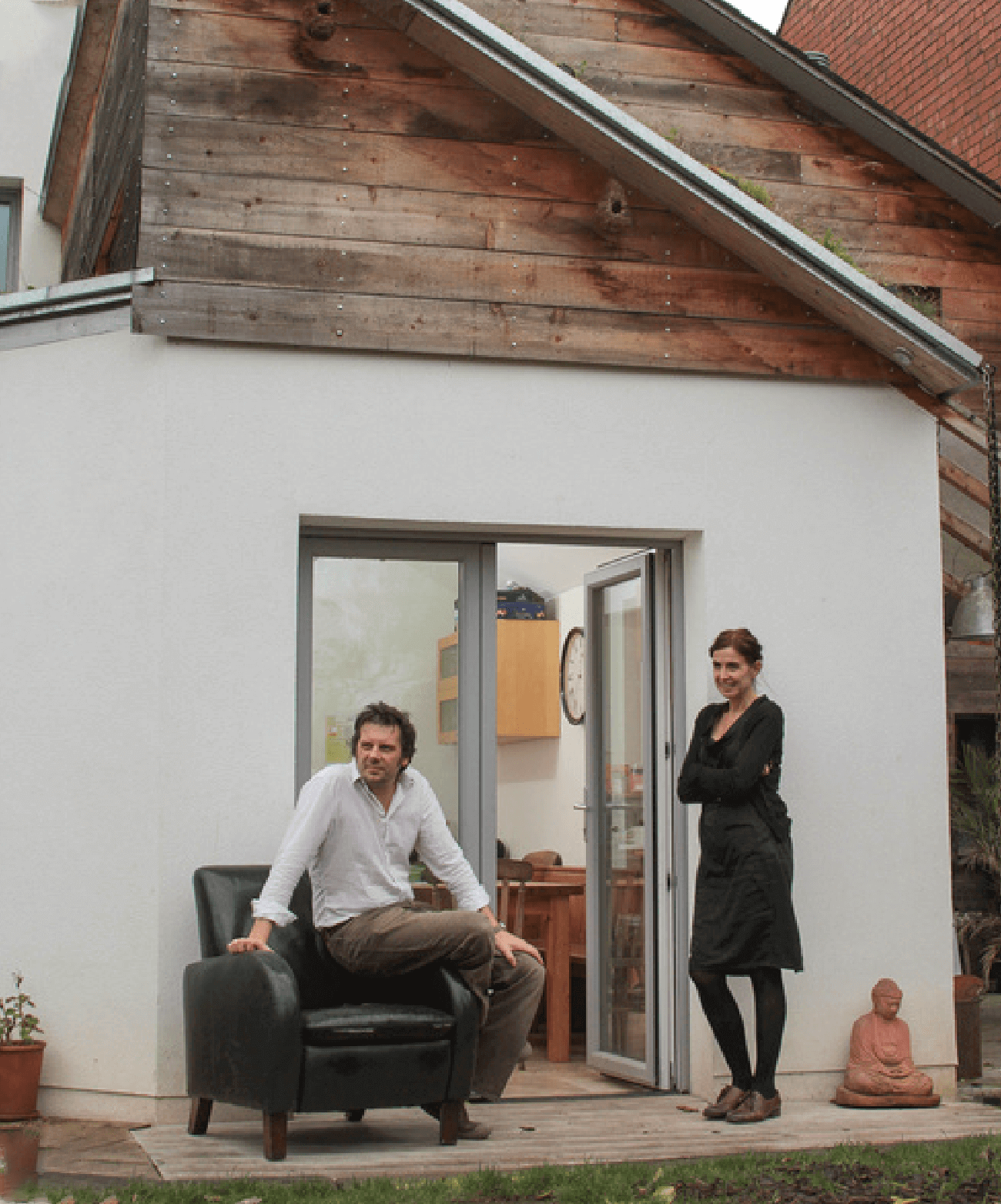 A man and woman stood outside an open door to a modern house. The man sat on an armchair, the woman smiling with arms folded looking relaxed