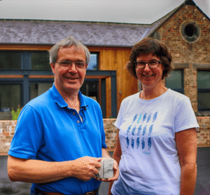 A man and woman wearing glasses holding a cup in front of a modern wooden and brick home with a circular window