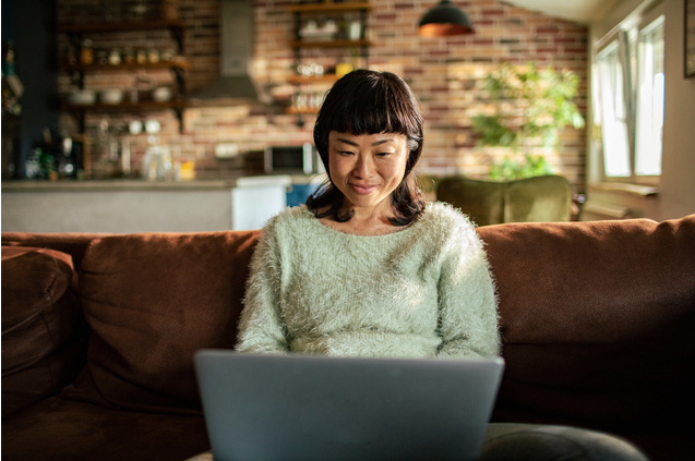 A woman in a green fluffy jumper sits on a brown sofa on her laptop in a modern, homely living room