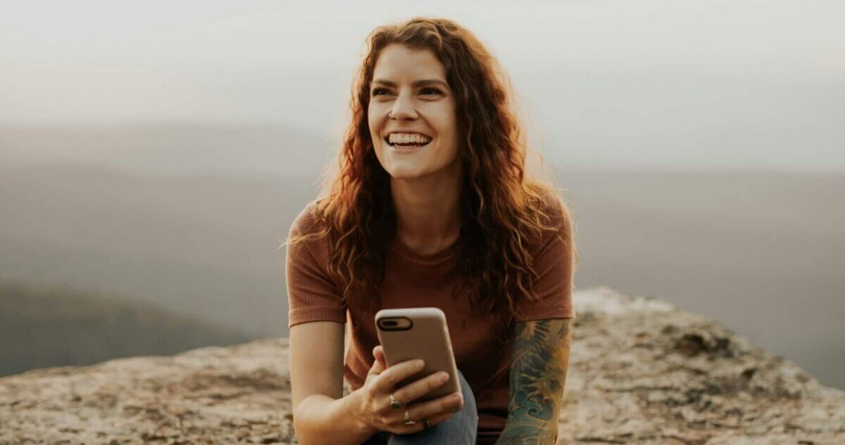 Young woman with red hair and a sleeve of tattoos on one arm, smiling sat on a hillside with a misty backdrop with her phone in her hand