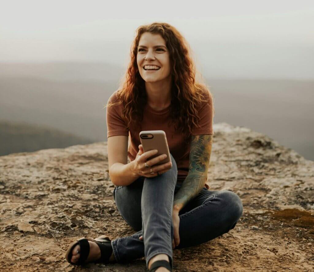 Young woman with red hair and a sleeve of tattoos on one arm, smiling sat on a hillside with a misty backdrop with her phone in her hand